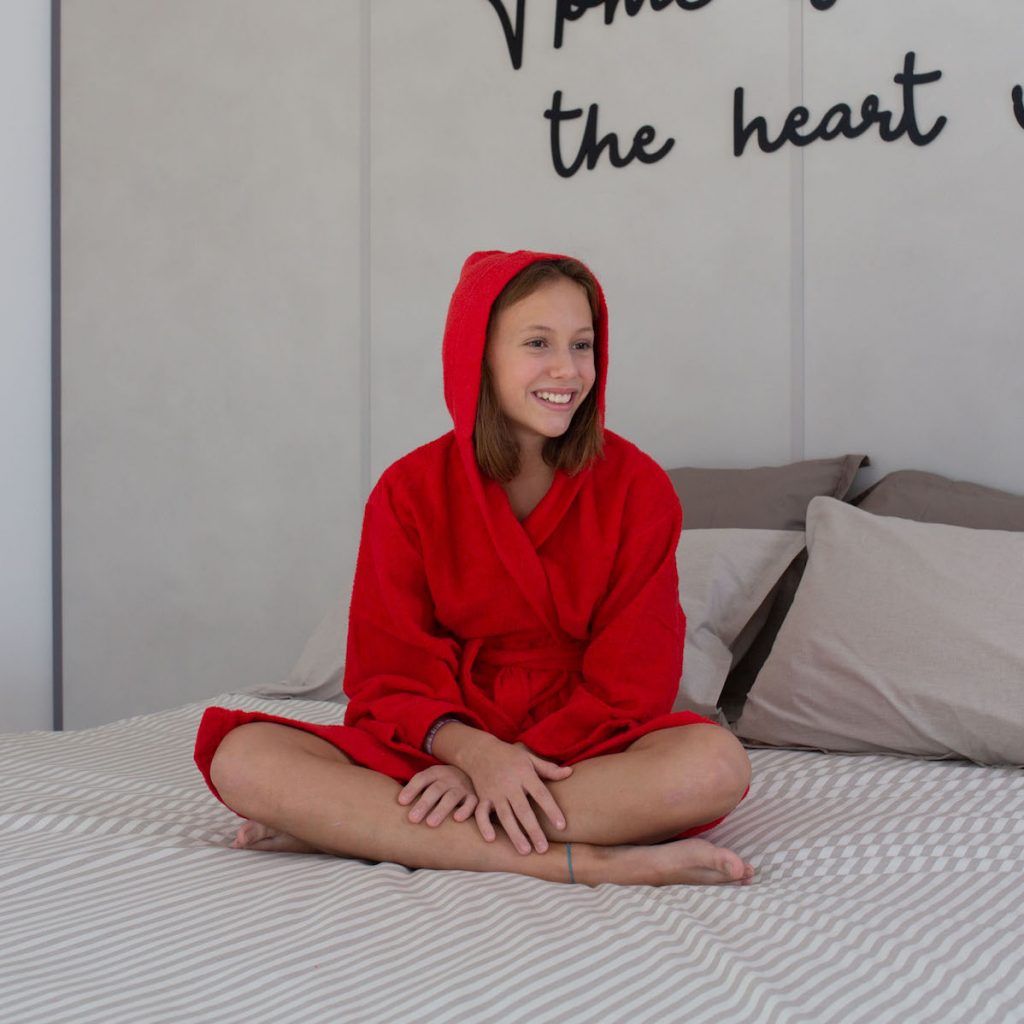accappatoio personalizzabile in spugna color rosso con bimba seduta sul letto che lo indossa